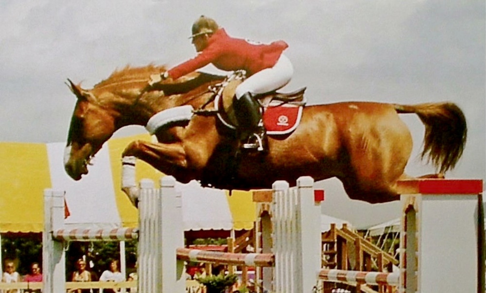 An old photo of a horse and rider jumping a fence.