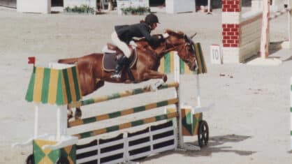 An old photo of a horse and rider jumping a fence in competition.