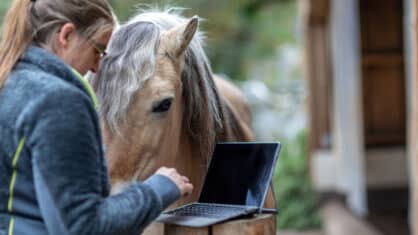 A woman typing info into a computer with a horse nearby.