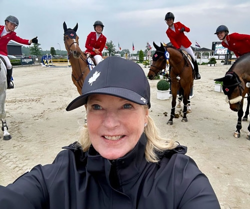 A woman taking a selfie with horses and riders behind.