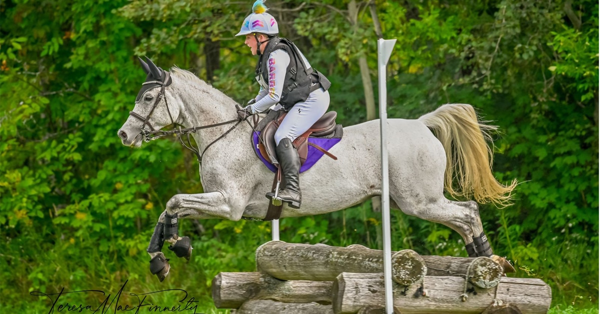 A woman on a grey horse jumping a cross-country fence.